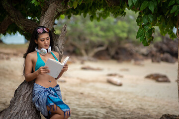 Young Woman Reading Book While Leaning On Tree At Beach