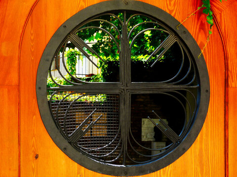 Round window opening in yellow stained pine wood exterior door with decorative metal grating. circular frame bolted the wood door. pine grain texture. green garden seen through the opening. lush green