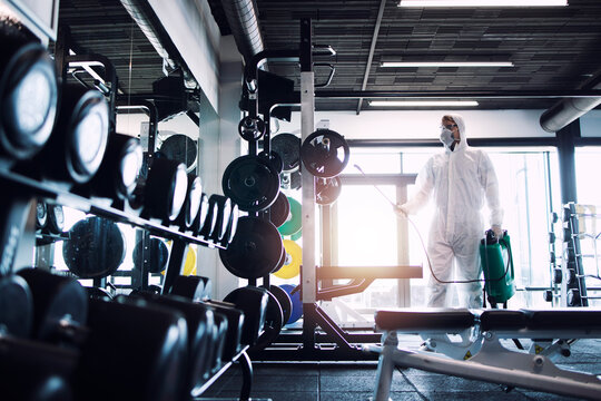 Gym disinfection and healthcare. Man in white protection suit disinfecting and fitness equipment and weights to stop spreading highly contagious coronavirus or COVID-19.