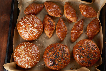 baked croissants and round buns with sesame seeds on an iron baking sheet with rolled brown baking paper