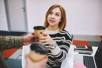 Charming blonde hipster student taking coffee beverage from colleague during break sitting at modern laptop computer in university.female designer holding cup with tasty drink enjoying free time