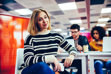 Portrait of charming blonde young woman dressed in casual outfit looking at camera while sitting in office interior.Attractive student with smartphone in hand during break in univerisity