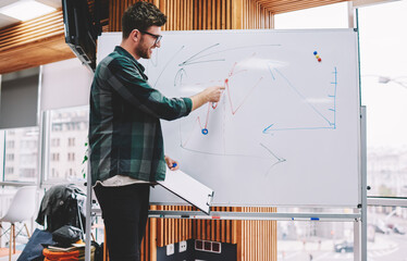 Professional male coach dressed in casual wear pointing on flip chart with graphics conducting...