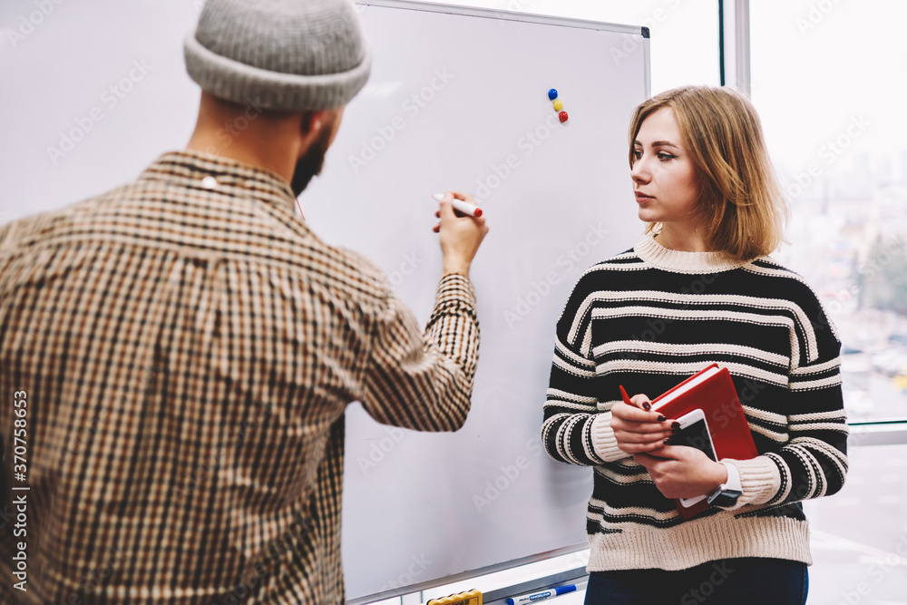 Poster hipster student writing examples of homework on board during training lesson with professional femal