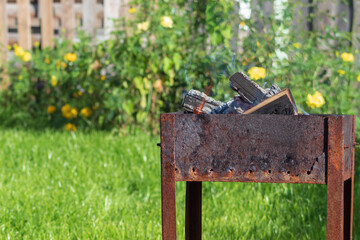 A worn-out barbecue with rust stands in the yard of the house against a background of flowers and grass. Wood burns in the grill