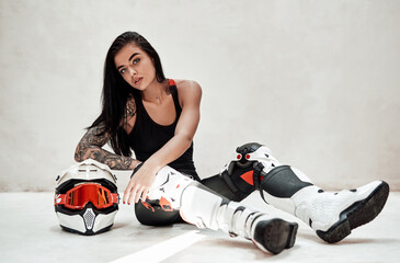 Fototapeta na wymiar Stylish looking brunette girl with tatoos in a black and red tracksuit, white boots and with a motorcycle helmet, posing in a white photo studio, sitting on the floor