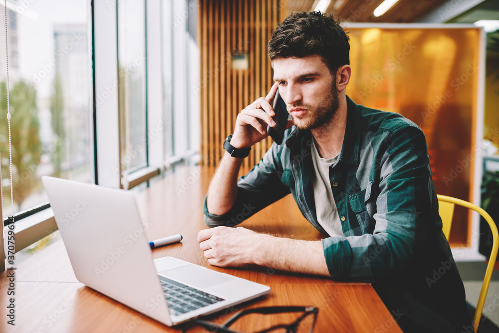 Poster young handsome male designer having mobile conversation via application on smartphone while watching