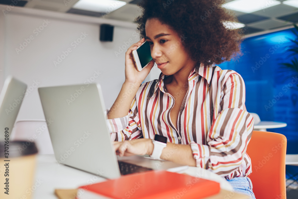Poster african american young woman calling on smartphone while typing information on laptop computer using