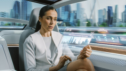 Attractive Senior Female Reading News on a Futuristic Transparent Tablet Computer with Augmented Reality Interface while Sitting on a Backseat of Autonomous Car. Self-Driving Van Rides on Public Road.