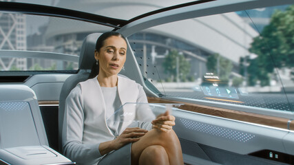 Attractive Senior Female Using a Futuristic Transparent Tablet Computer with Augmented Reality Interface while Sitting on a Backseat of Autonomous Car. Self-Driving Van Rides on Public Road.