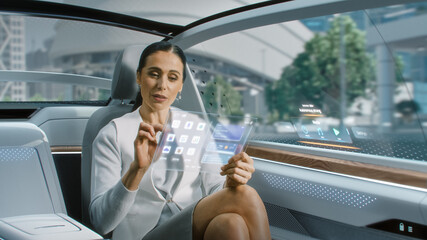 Attractive Senior Female Reading News on a Futuristic Transparent Tablet Computer with Augmented Reality Interface while Sitting on a Backseat of Autonomous Car. Self-Driving Van Rides on Public Road.