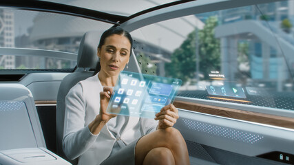 Attractive Senior Female Reading News on a Futuristic Transparent Tablet Computer with Augmented Reality Interface while Sitting on a Backseat of Autonomous Car. Self-Driving Van Rides on Public Road.