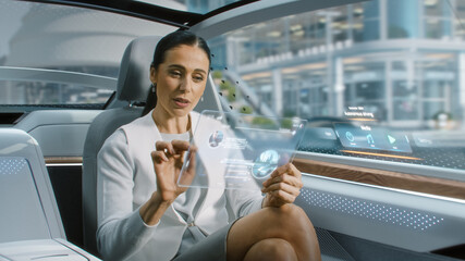Attractive Senior Female Reading News on a Futuristic Transparent Tablet Computer with Augmented Reality Interface while Sitting on a Backseat of Autonomous Car. Self-Driving Van Rides on Public Road.