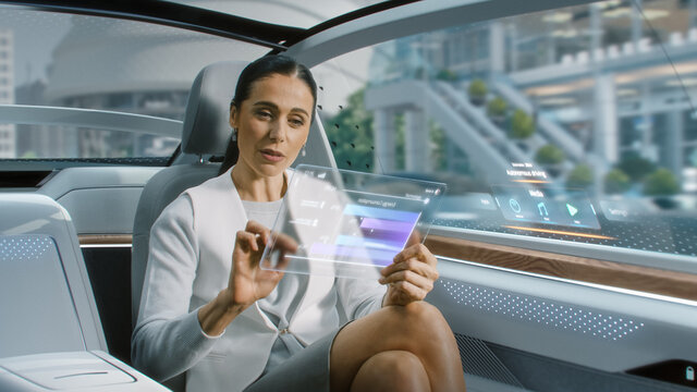 Attractive Senior Female Reading News on a Futuristic Transparent Tablet Computer with Augmented Reality Interface while Sitting on a Backseat of Autonomous Car. Self-Driving Van Rides on Public Road. - Powered by Adobe