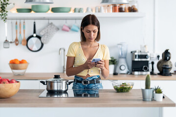 Concentrated young woman using her mobile phone while eating a salad in the kitchen at home.