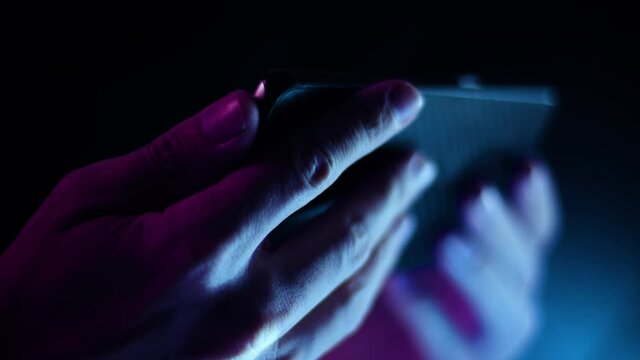 Thumb scrolling on a smartphone in landscape close up in a dark room with purple and blue lighting soft focus