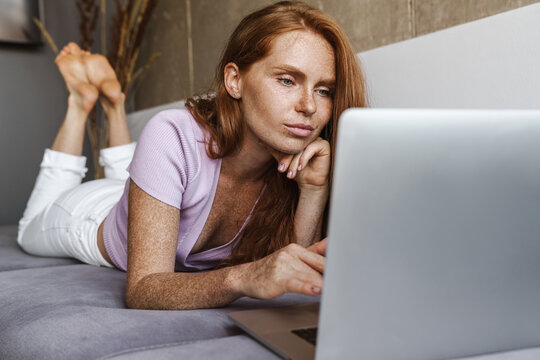 Image of redhead concentrated woman using laptop while lying on couch