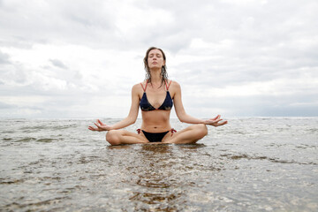 Fototapeta na wymiar Woman in yoga meditation pose on the beach. Young healthy woman practises yoga in the shallow ocean water with blue sky behind.