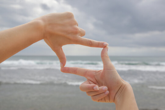 Woman holds hands in the shape of a frame in front of blue ocean horizon