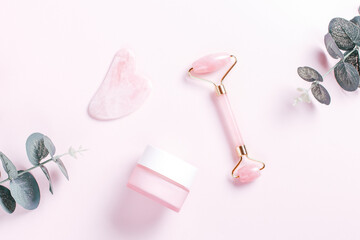 Facial roller, gua sha and moisturizing cream with green leaves. Natural cosmetics and facial tools on pink. Rose quartz beauty roller, heart gua sha flat lay. Tools for skin care routines, top view