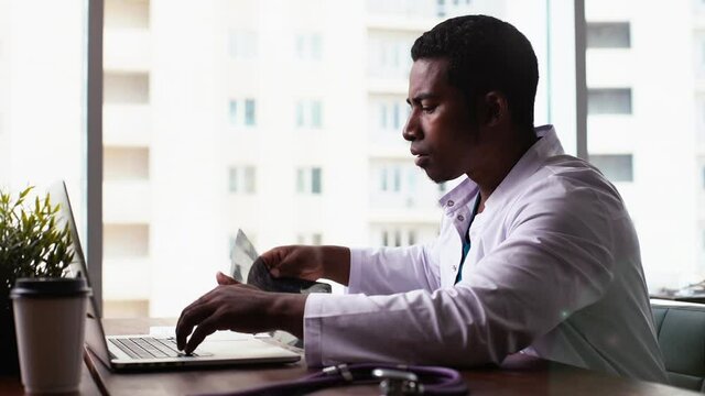 Serious African American black man doctor in white coat uniform working typing on laptop computer, looking at MRI scan of head, thinking on diagnosis, sitting at desk on workplace at hospital. 