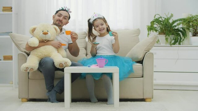 Father, daughter and teddy have tea party. They are sitting on the sofa wearing crowns, offering you to taste some sweet tea.