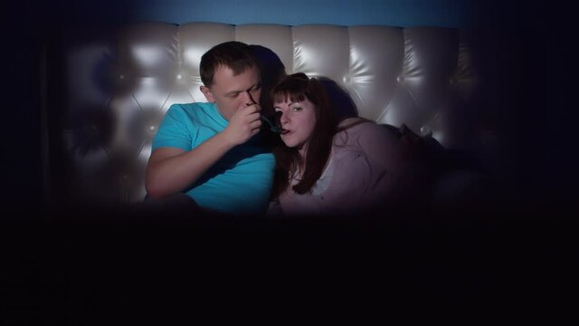 Night, a couple lies on the bed in front of the TV, a man feeds a woman with ice cream and eats himself, camera movement