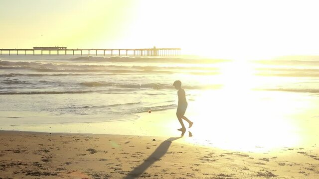 Child Silhouette Playing With Ball at Shoreline During Bright Golden Sunset Near Pier