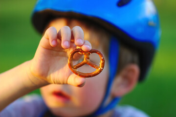 A boy with a bicycle helmet holds a small pretzel in his hand. Break snacks.
