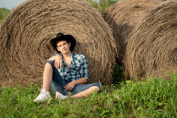 Naklejka na ściany i meble A young man in a plaid shirt, a cowboy hat with a straw in his mouth sits near a roll of hay and looks into the distance. Farmer man sitting by the hay.