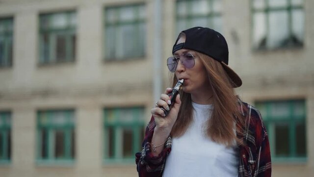 Close up pretty young hipster woman in black cap vape ecig. Girl vape popular ecig gadget, vaping device.Happy brunette vaper girl with e-cig. Portrait of smoker female model with electronic cigarette