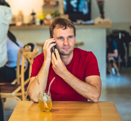 Restrained smile happy young man answer smart phone in red shirt in cafe drinking ice tea, waiting somebody or coworking. Real people expression, life problems solving or communication, relationship