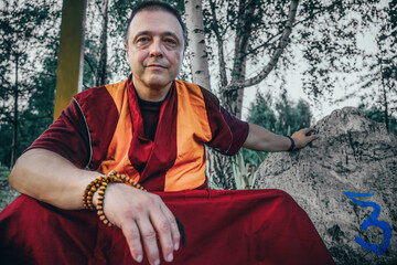 lama monk with a wooden rosary around his neck chides near a large stone