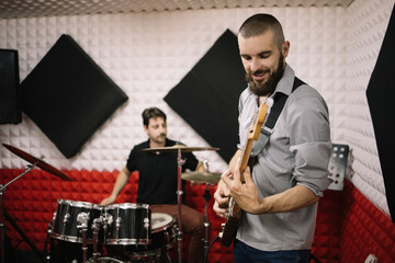 Music band playing instruments at soundproof studio. Two band members playing electric guitar and drummer set and recording.
