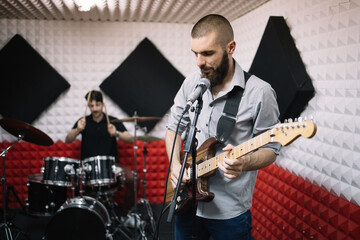 Rock band working in music recording studio. Man singing while playing the guitar in music studio with blurred drummer on drum set.