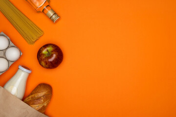 Bread and bottle of milk in paper bag. Spaghetti, sunflower oil, eggs and red apple on orange background. Healthy food, delivery, donation concept. Food stock for quarantine. Top view, copy space
