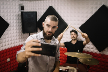 Bearded man taking selfie with musician who is holding drumsticks. Young man is photographing himself and blurred drummer.