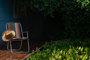 A summer hat and a folding chair surrounded by green vine leaves on a wooden deck. Cottage...