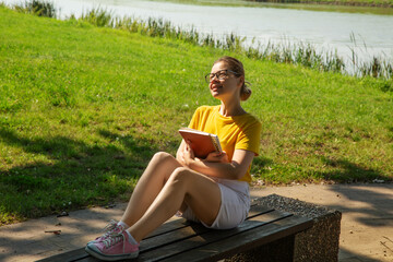  Beautiful female college student in glasses holds books and smiles in a park outdoors sitting on a bench