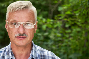 Close-up portrait of handsome gray-haired adult man with mustache and glasses smiling on the background of a summer forest park