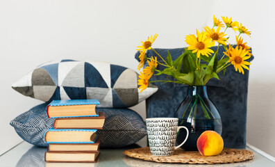 Cozy home interior decor: stack of books, peach, cup of coffe, decorative pillows, box with plaid and vase with yellow flowers on a table. Distance home education.Quarantine concept of stay home.