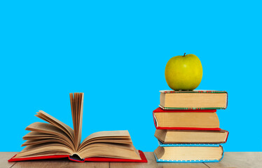 Open book textbook, pile of books and green apple on a wooden desk on the blue background. Back to school distance home education.Quarantine concept of stay home.