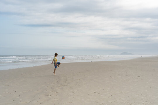 Menino, brasileiro, usando camiseta e bermuda, jugando futebol sozinho na praia de Guaratuba, litoral norte de São 
Paulo. 