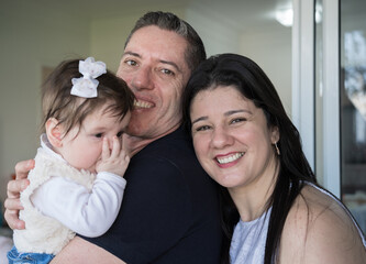 Obraz premium Homem sorridente segurando uma criança no colo ao lado de sua esposa sorrindo. Família brasileira. Padrinhos com sua afilhada no colo. 