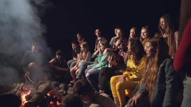 Kids singing and playing guitar near camp fire.
