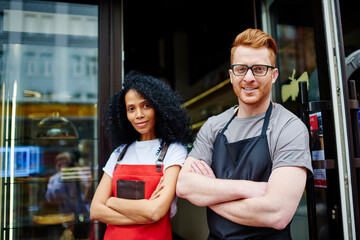 Portrait of prosperous team of two multicultural professional barista and waitress with crossed hands and aprons smiling at camera while inviting clients in own cafeteria standing outdoors