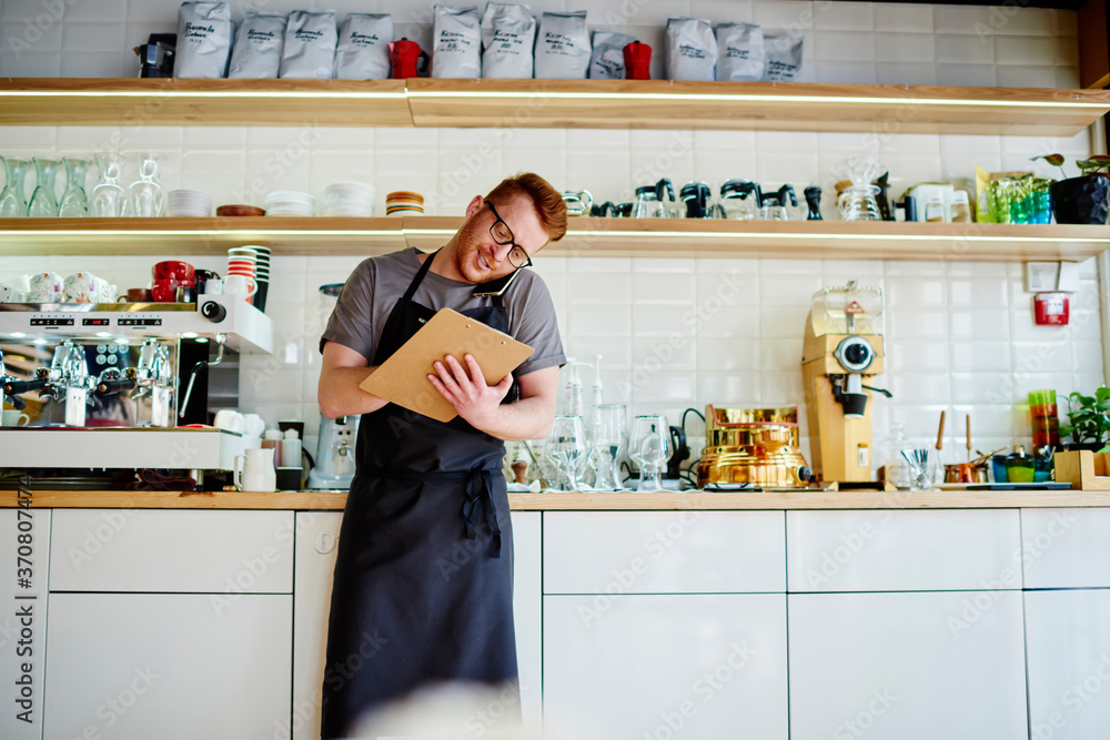 Poster caucasian barista calling on smartphone device connected to wireless 4g internet and checking order 