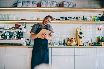 Caucasian male owner of coffee shop in black apron standing against bar with machine and calling on smartphone to make product order for development business.Employee of restaurant talking on cellular