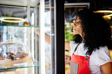 African american young woman confectioner in red apron taking tasty chocolate cake from candy refrigerator in own cafeteria interior.Dark skinned female waiter opening frog with delicious pastry