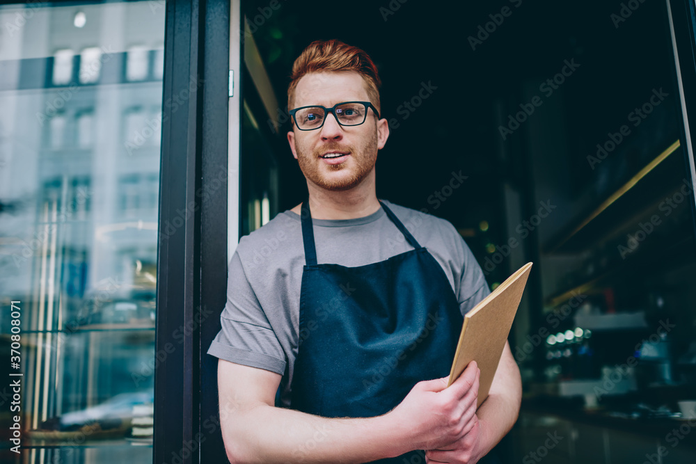 Poster professional caucasian waiter in black apron holding menu and smiling while inviting guests to own b